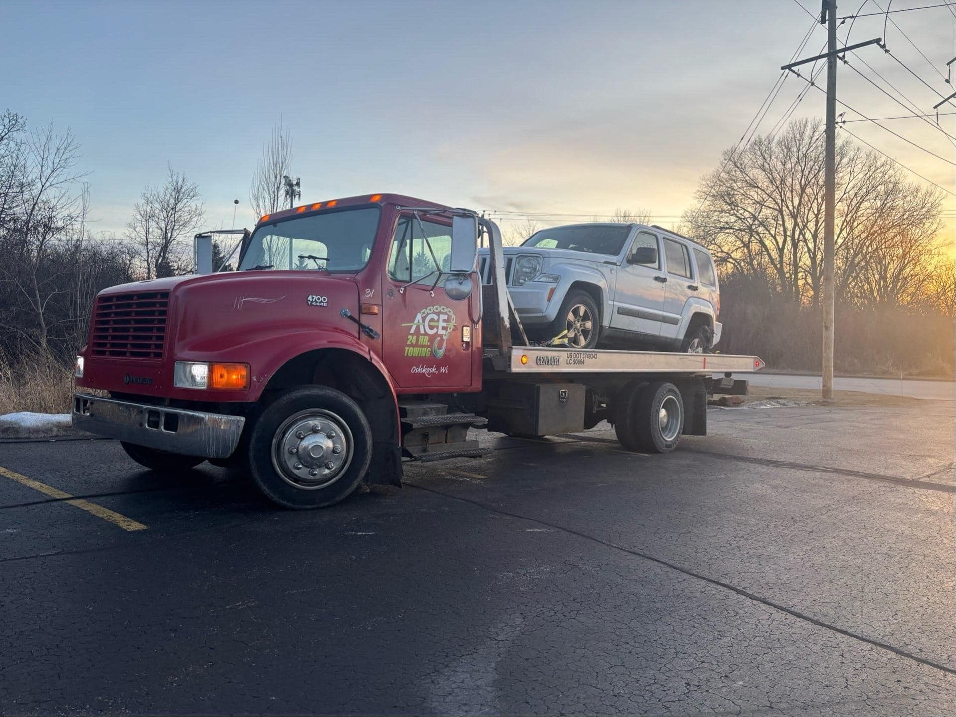 Red tow truck carrying a white Jeep on a road near a field at sunset.