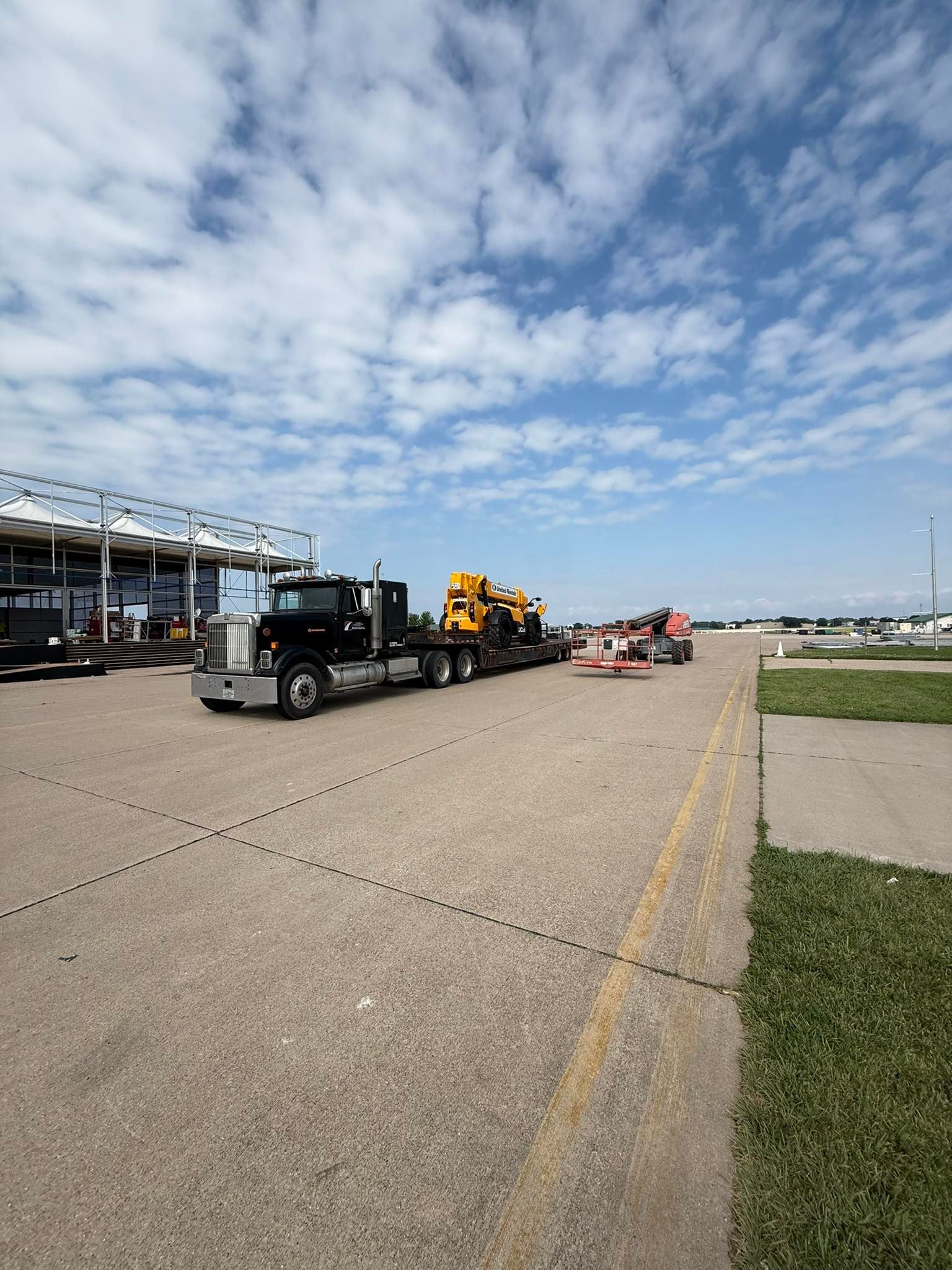 Black semi-truck hauling yellow machinery on a flatbed trailer, outdoors on a sunny day.