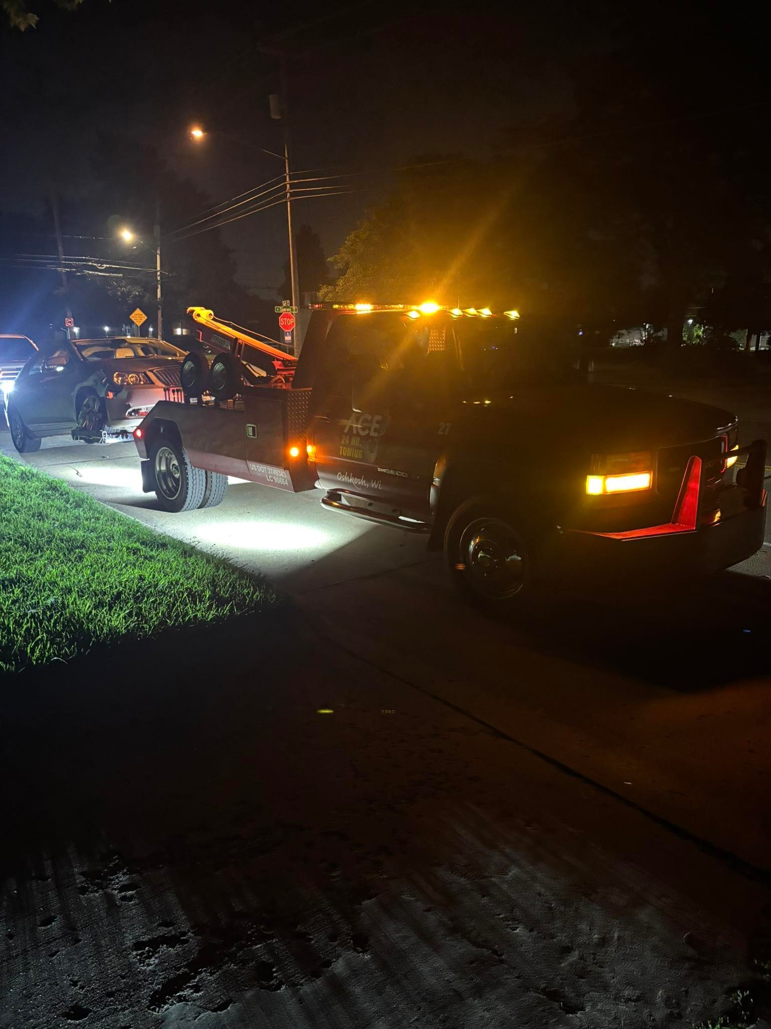 Tow truck at night with flashing lights towing a car on a dark road, beside a grassy area.