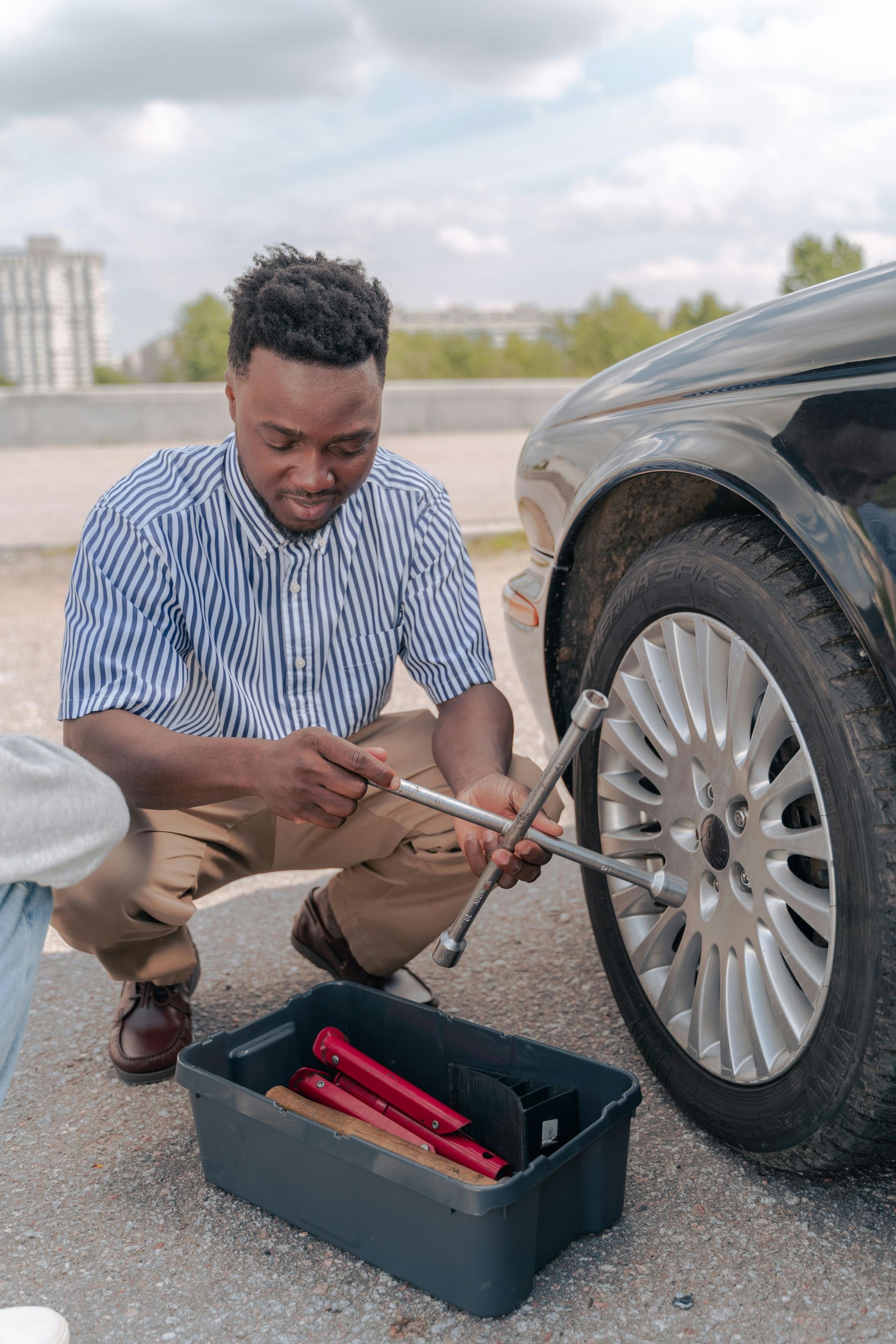 Man changing a tire on a car; using a wrench; tools in a box on the asphalt.