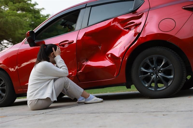Woman sits near a damaged red car, looking distraught. Car has a large dent in the side.