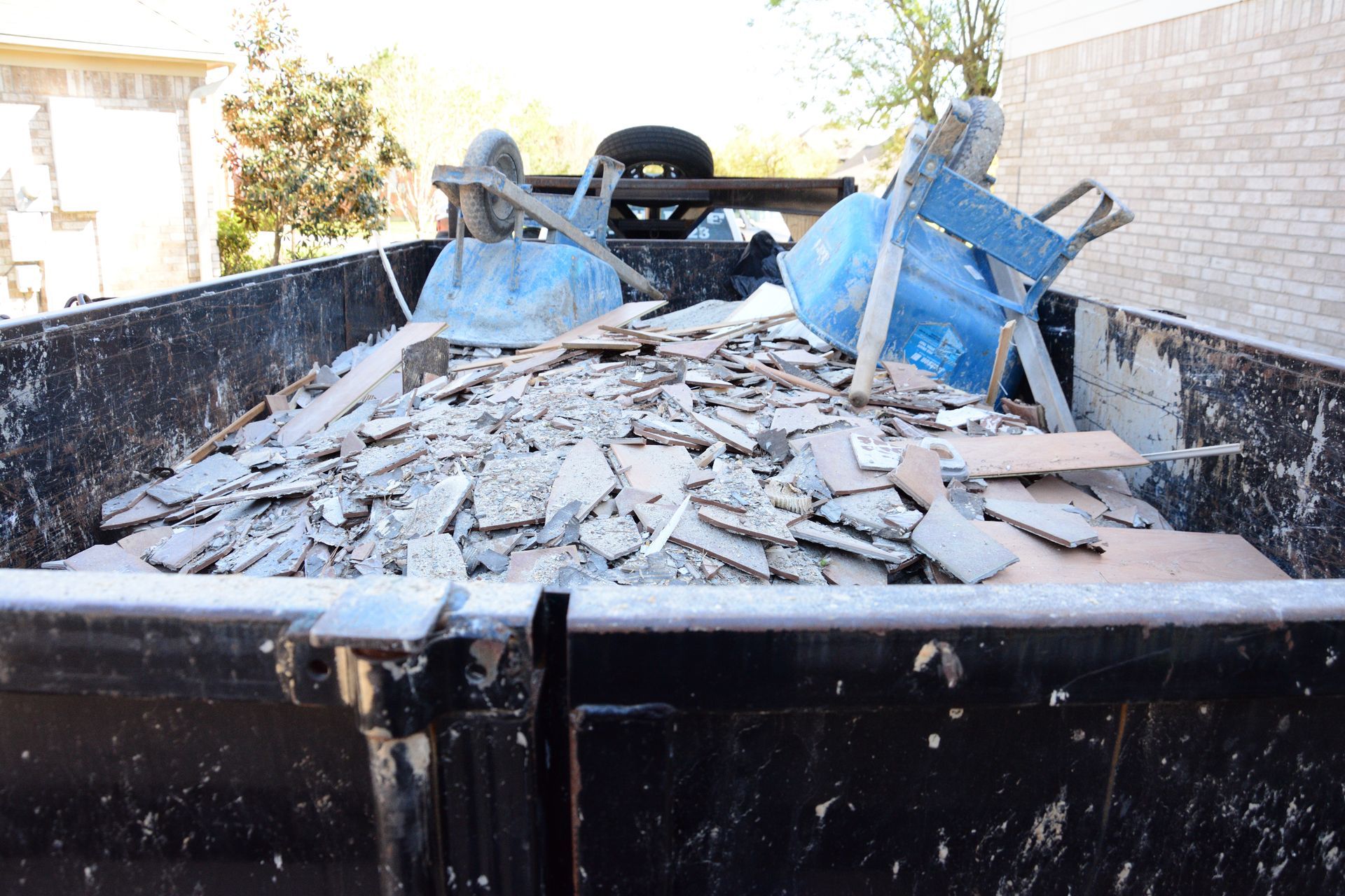Dumpster filled with construction debris, including broken tiles and a blue chair. Outdoors, daytime.