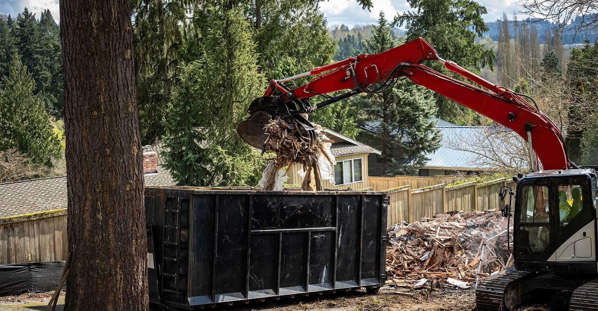 Red excavator loading debris into a large black dumpster near a house.