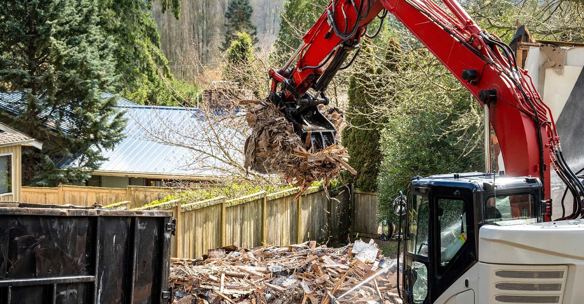 Red excavator demolishing a wooden fence, debris in a metal container, trees in the background.