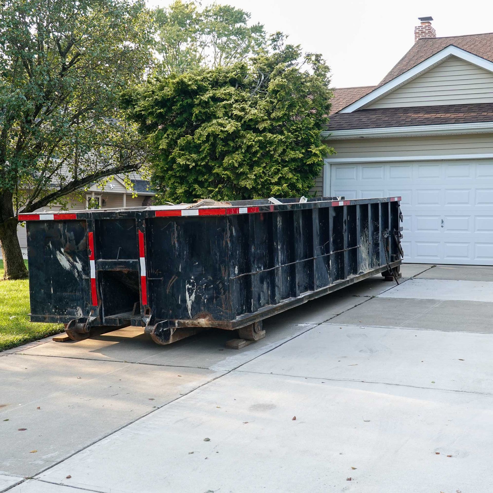 Black dumpster on a driveway in front of a house, near a tree.