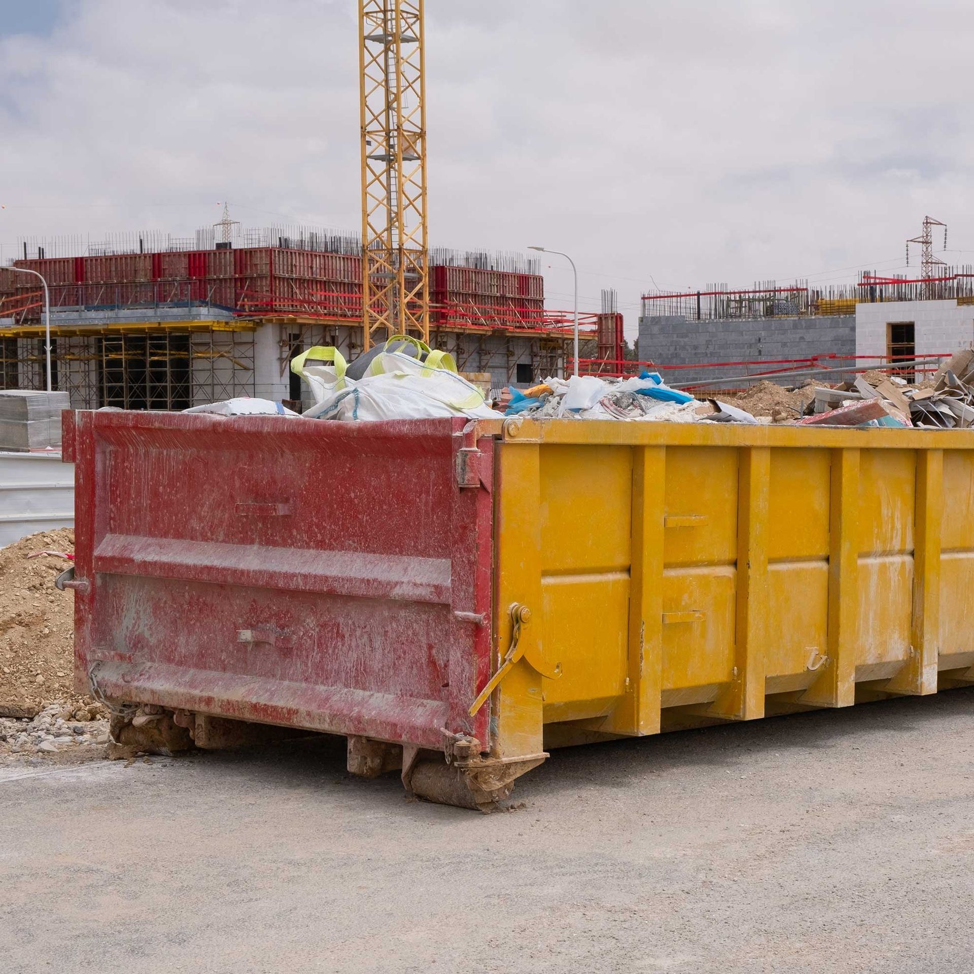 Red and yellow dumpster overflowing with construction debris at a building site with a crane in the background.