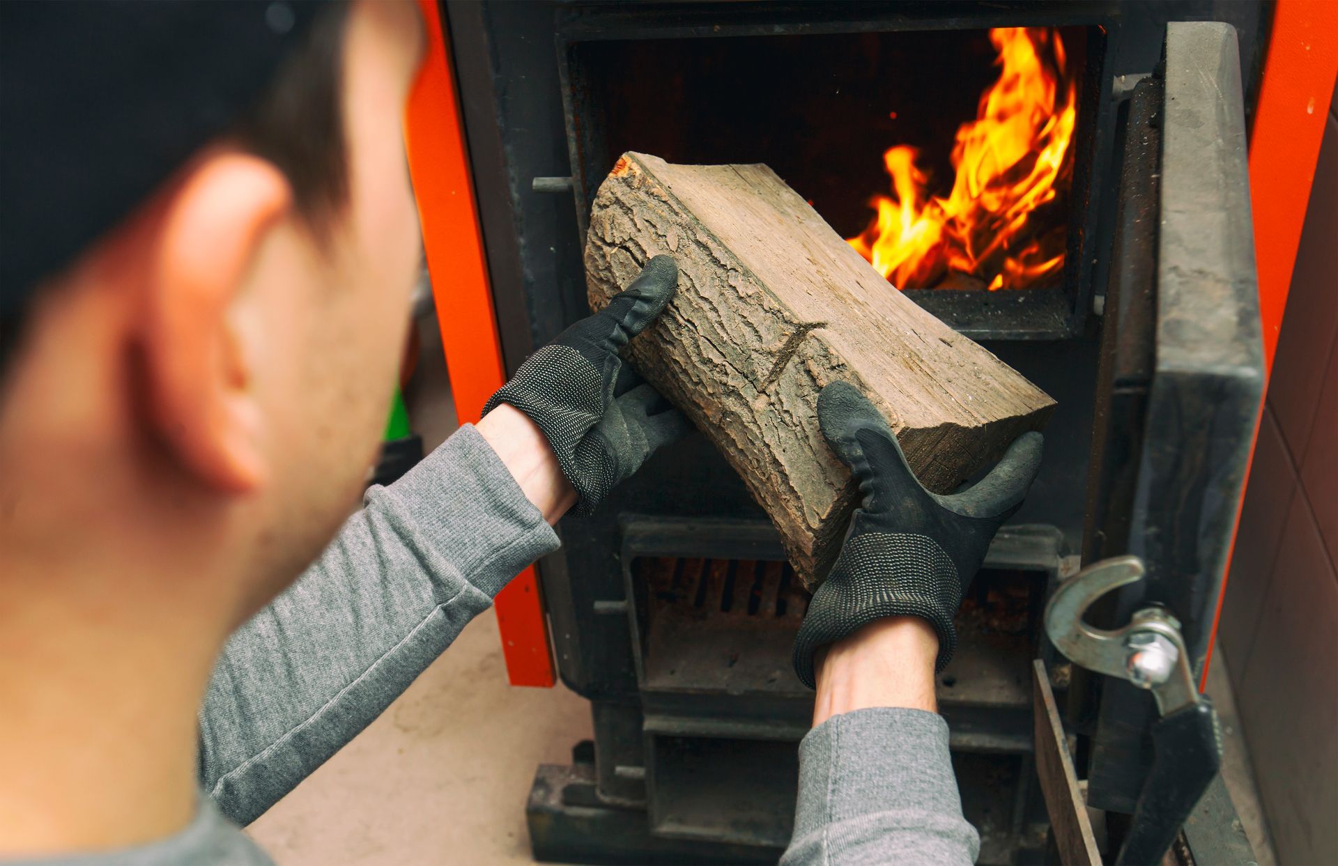 Person adding a log to a wood-burning stove with flames. Gloves on, gray shirt, orange and black stove.