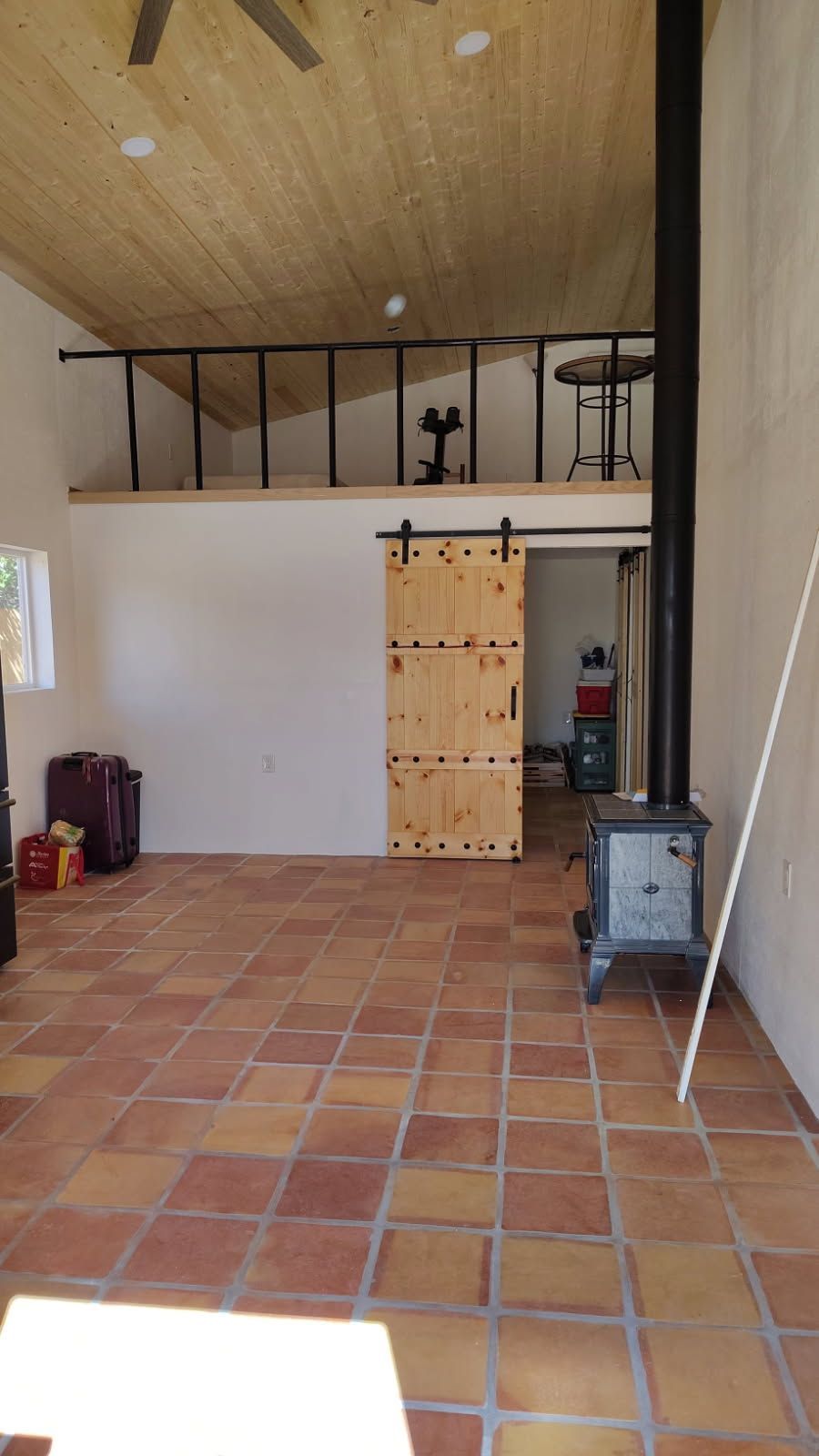 Interior with terracotta tile floor, loft, barn door, wood-burning stove, and black metal railing.