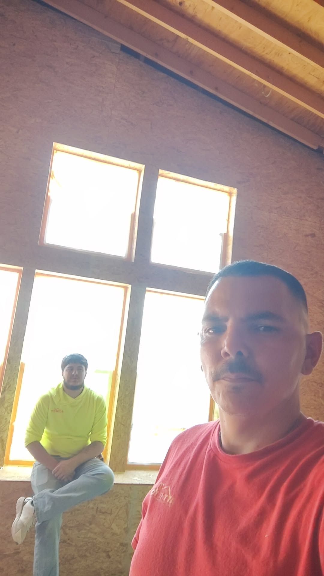 Two men in a building under construction; one in red shirt takes a selfie. The other sits near a window.