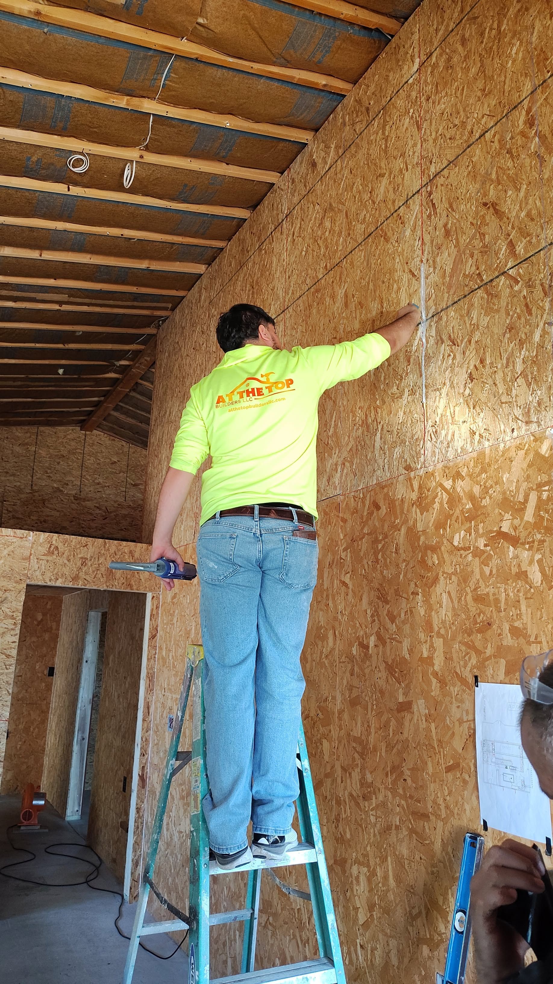 A construction worker in a neon shirt on a ladder installs wiring on an OSB wall.