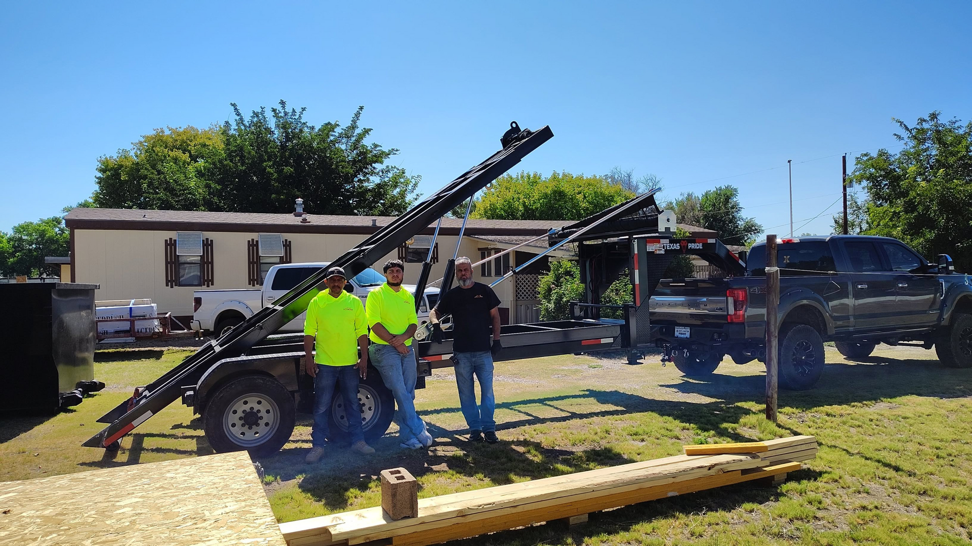 Three men beside a trailer loaded with long, dark beams, parked next to a truck, house, and wood on a sunny day.