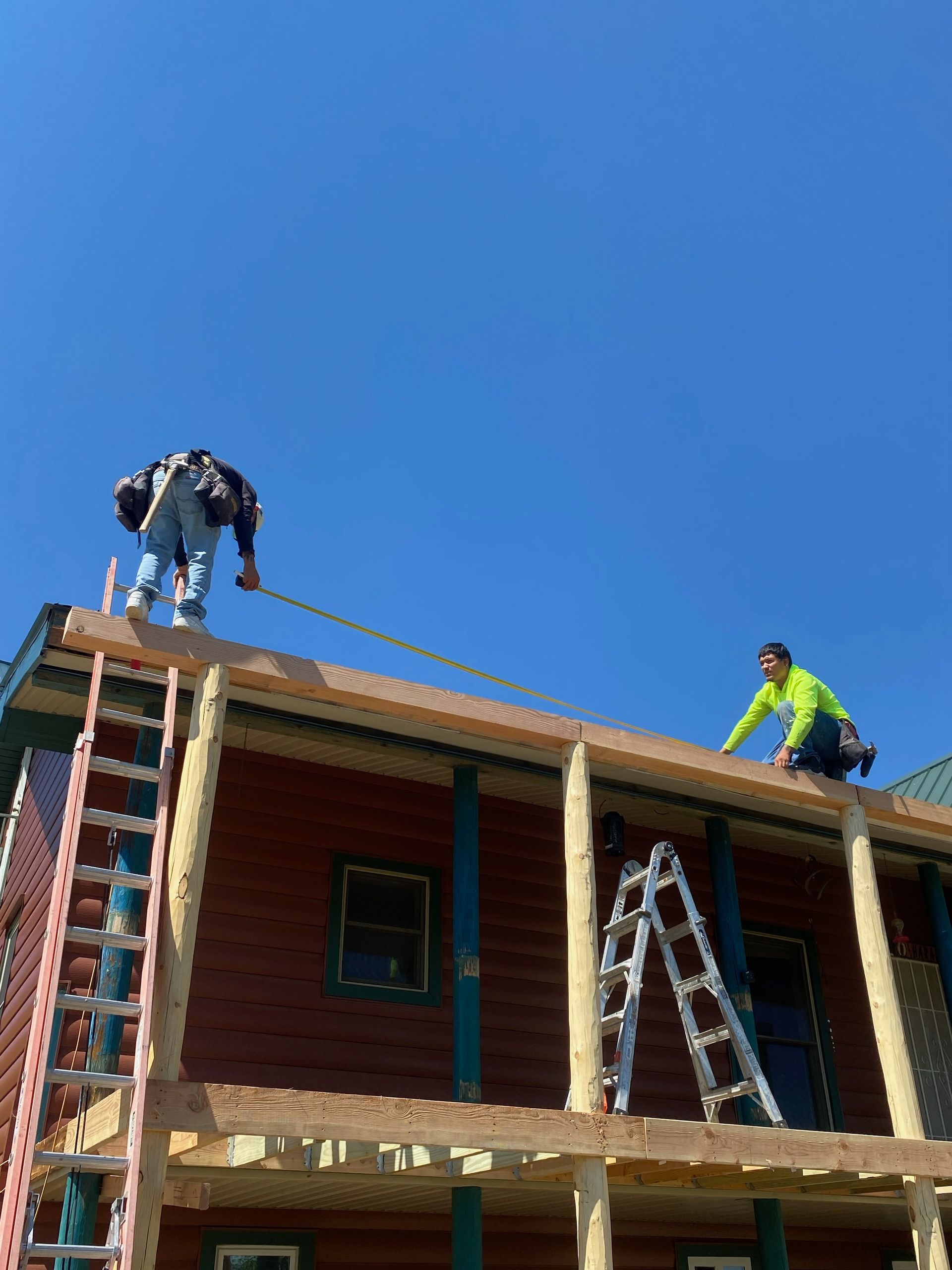 Two workers on a building roof, measuring, on a sunny day. One stands, the other kneels.
