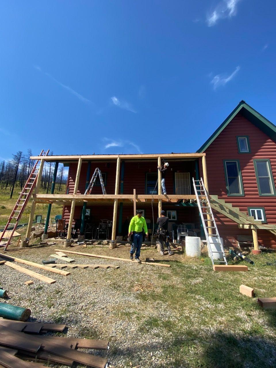 Construction of a wooden deck on a red house under a clear blue sky; a worker stands in front of the house.