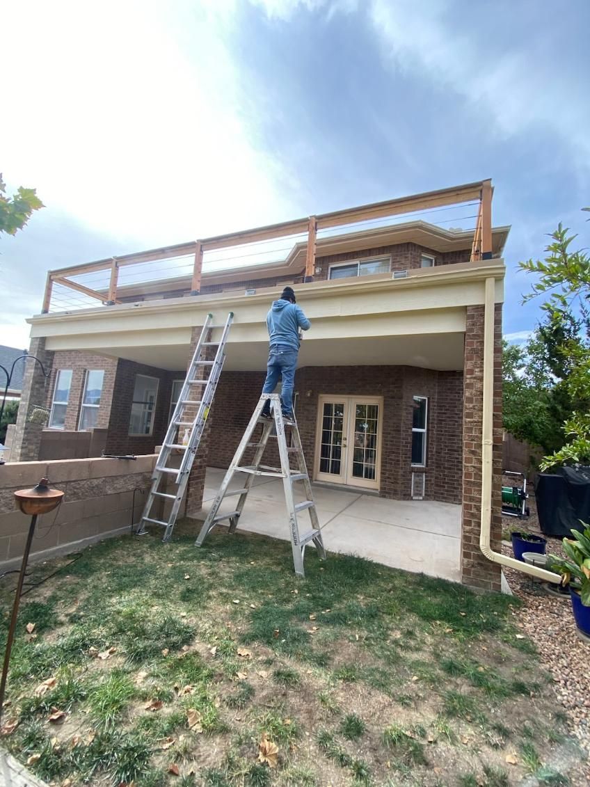 A person on a ladder working on the wooden railing of a brick house patio.