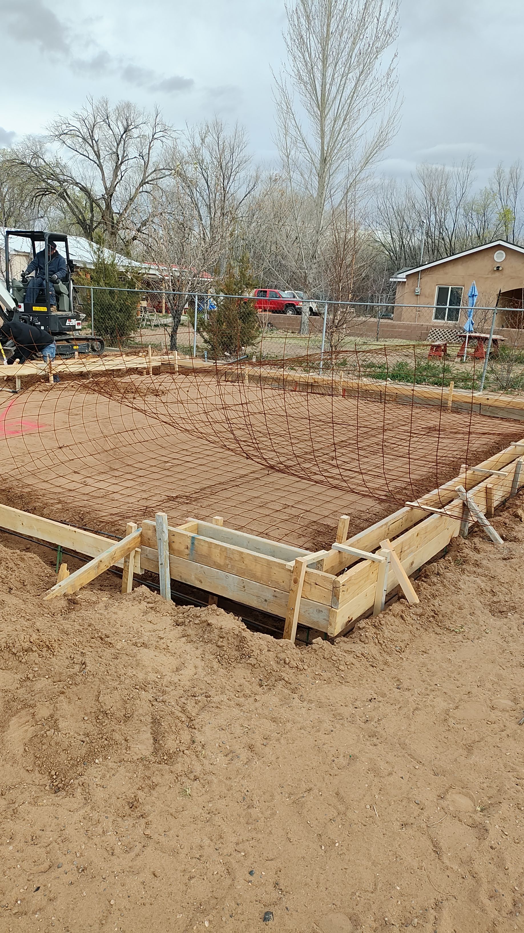 Construction site with wooden forms and dirt, likely for a foundation. An excavator is in the background.