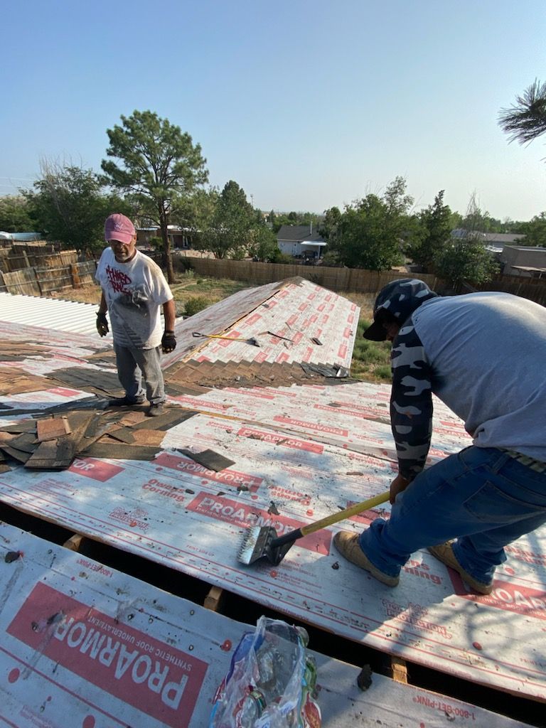 Two roofers on a rooftop, removing shingles. One uses a shovel, the other stands, overseeing. Daytime, sunny.