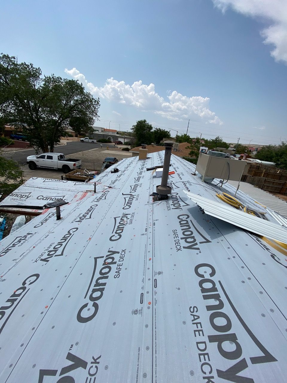 Rooftop with new roofing material installed. Sky is blue with clouds. Cars and trees are in the background.