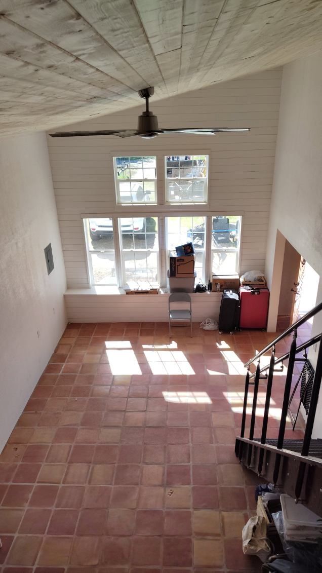 High-ceilinged room with tiled floor, large windows, and a ceiling fan. Sunlight streams in.