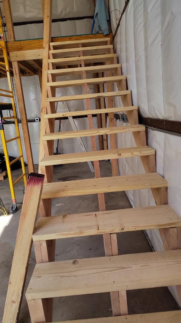 Wooden staircase in a building. Steps are unfinished; the setting is indoors with wall panels and a scaffold.