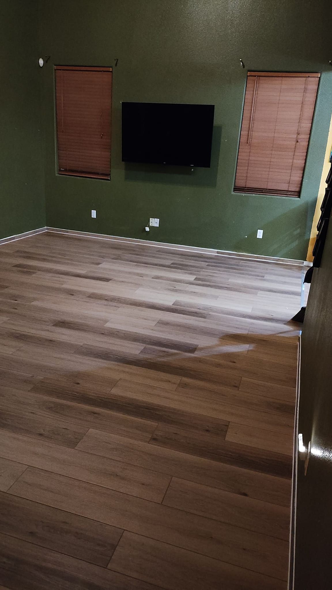 Green-walled room with wood-look floor; TV on wall, blinds on windows.