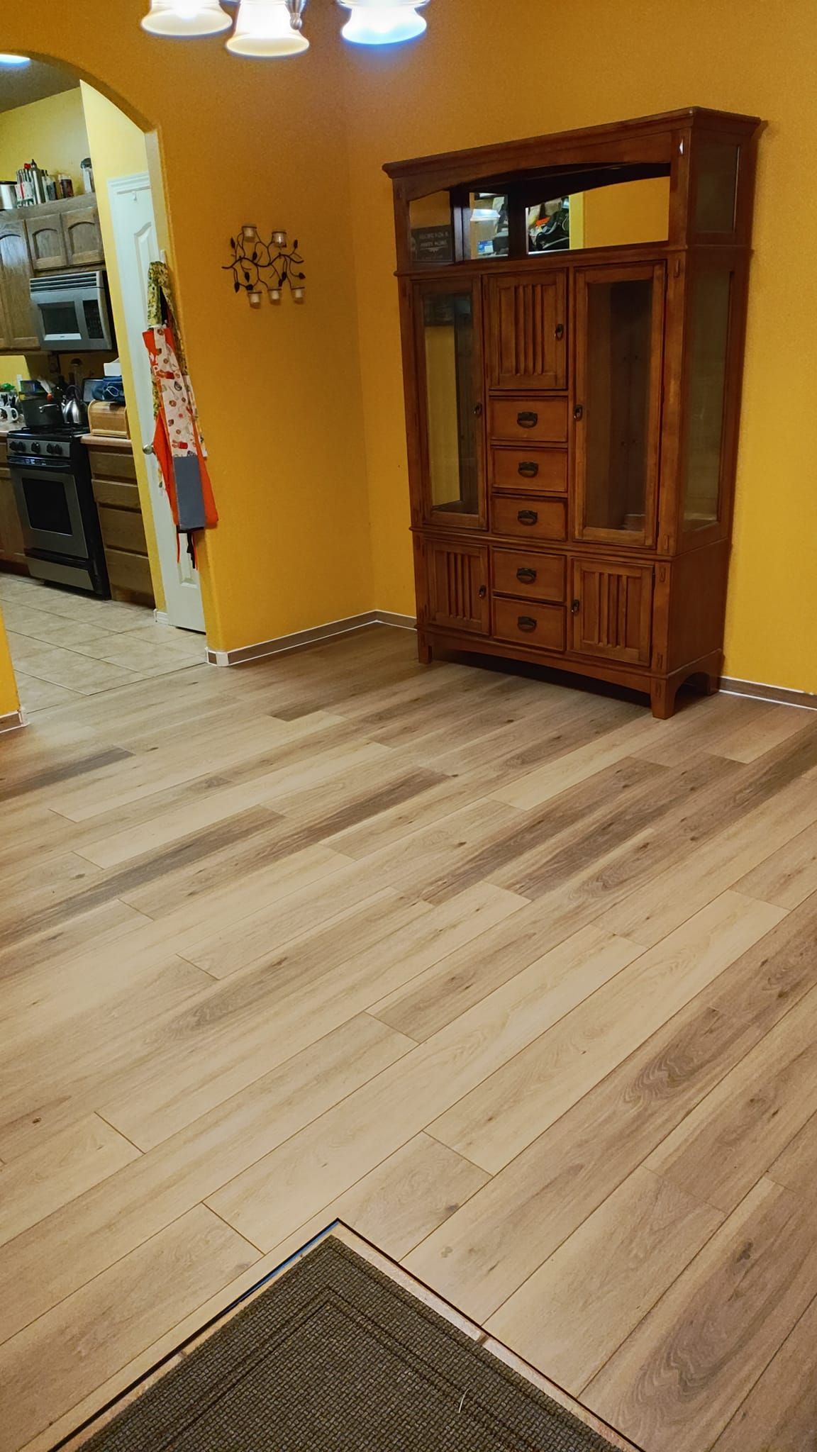 Wooden cabinet in a room with light wood floor and yellow walls, next to a doorway to a kitchen.