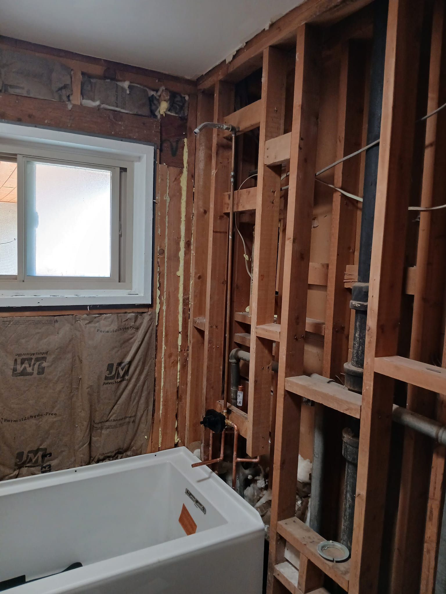 Bathroom under renovation: exposed studs, bathtub, window, plumbing visible. Brown, white tones.