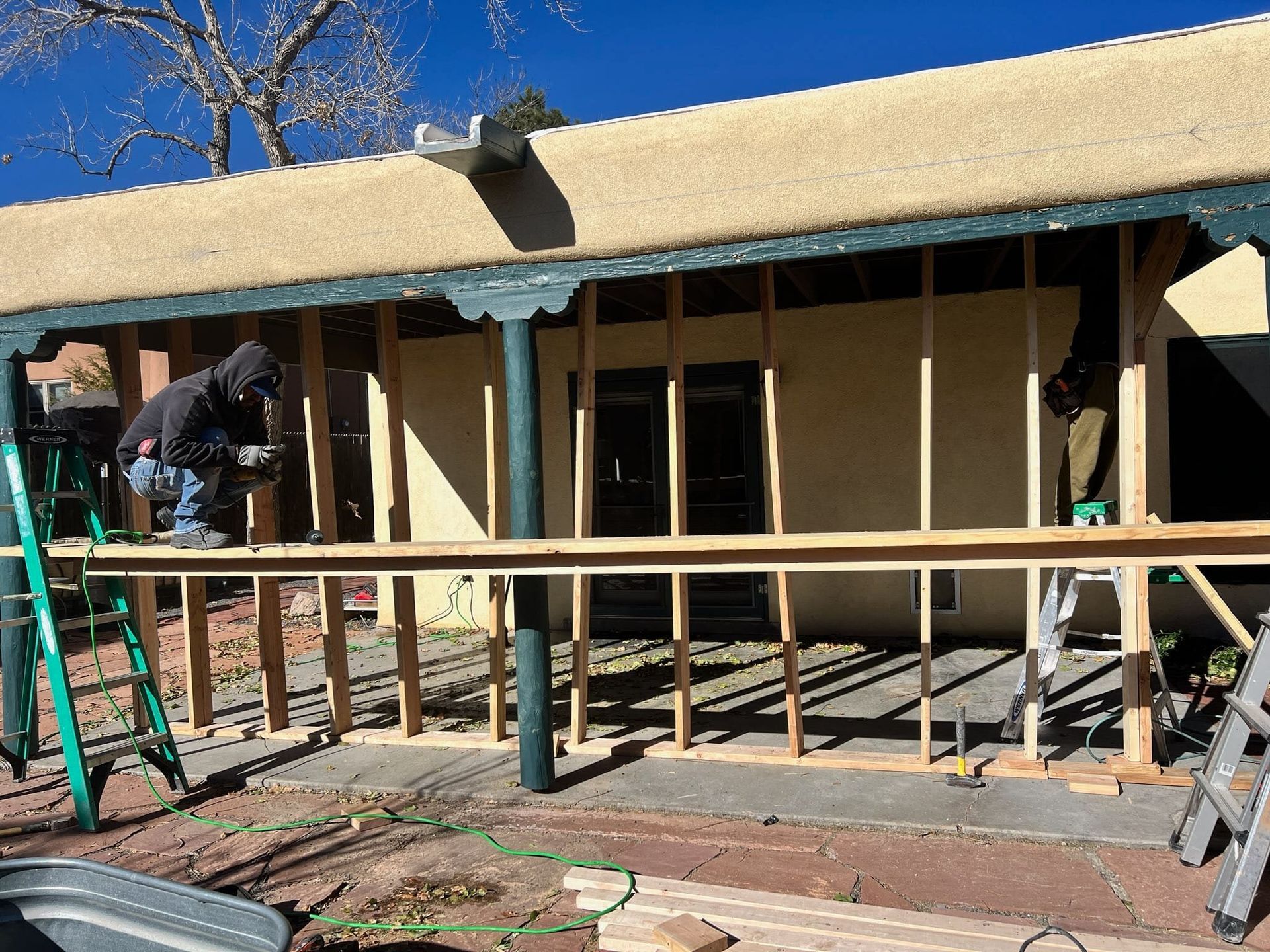 Construction workers building an outdoor structure with wooden framing, in front of a tan building, on a sunny day.