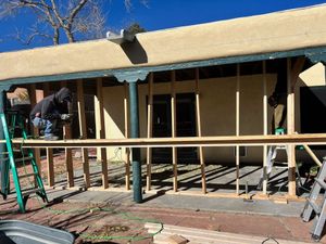 Construction workers building an outdoor structure with wooden framing, in front of a tan building, on a sunny day.