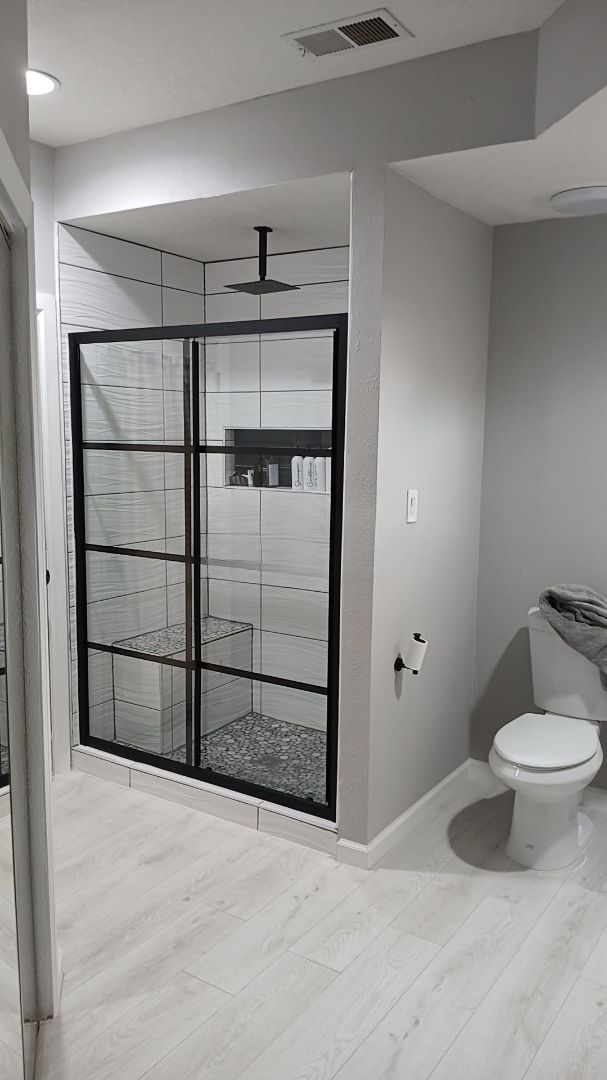 Bathroom with modern shower, black-framed glass door, pebble floor, light wood-look tile, and white toilet.