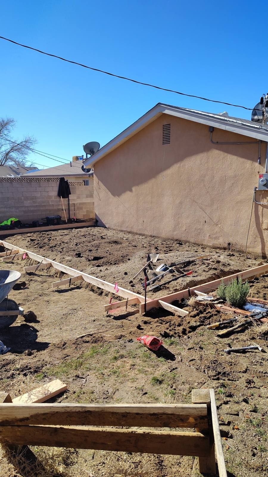 Backyard construction site: wooden forms outline a future concrete slab. Brown dirt, beige house, and clear blue sky.