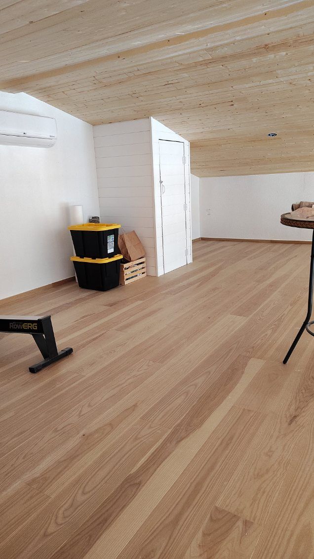Wooden-floored room with white walls, wood-paneled ceiling, storage containers, and a small table.