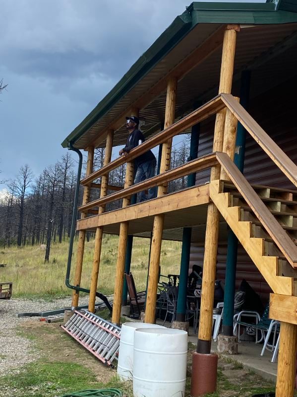 Person on wooden deck of cabin with stairs. Overcast day, green roof, and barrels in front.