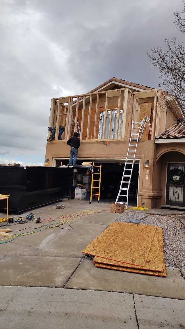 Man on ladder building addition to a two-story house. Garage door open; cloudy sky.
