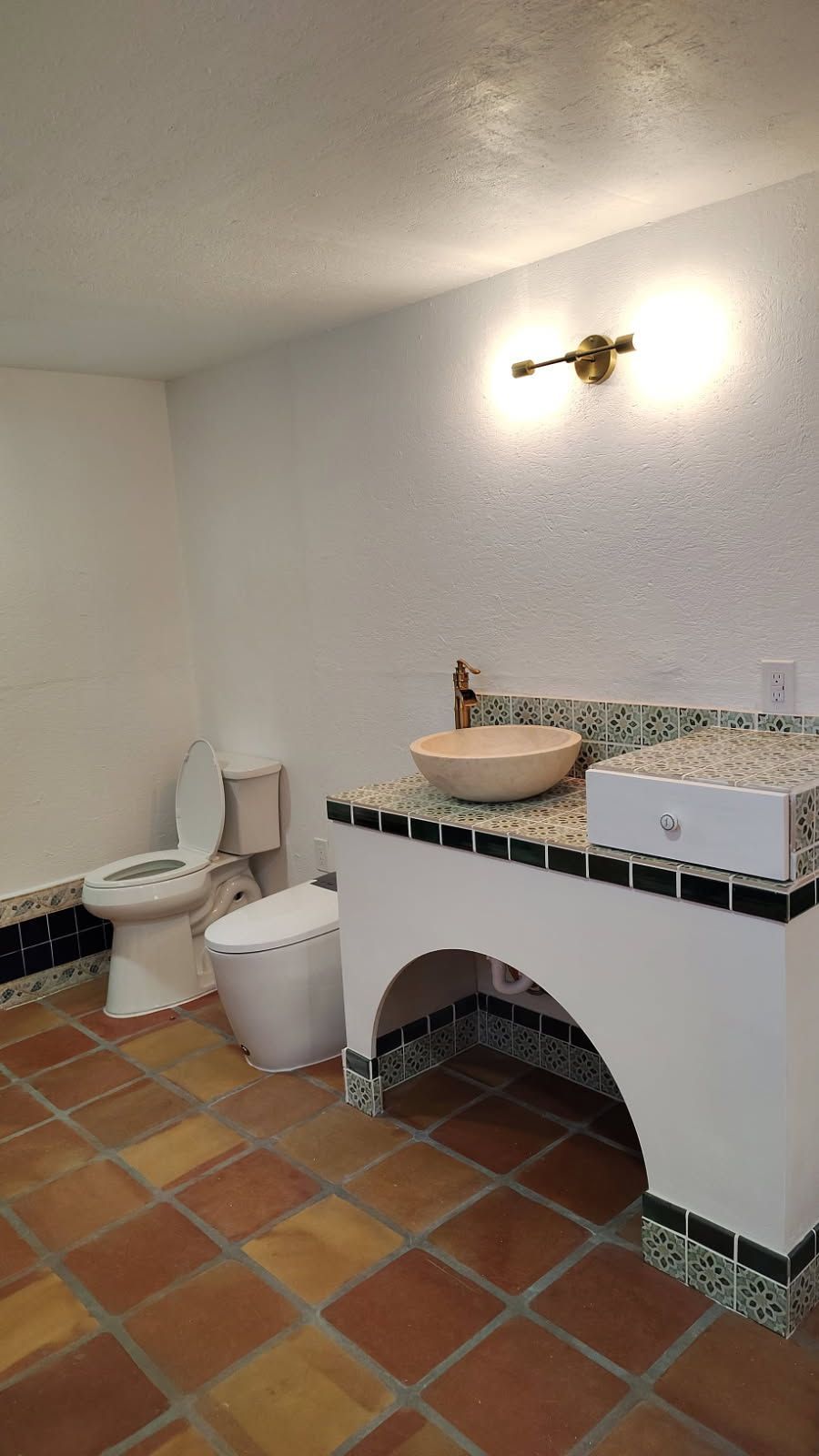 Bathroom with terracotta tile floor, white walls, and a decorative sink. Toilet is visible.