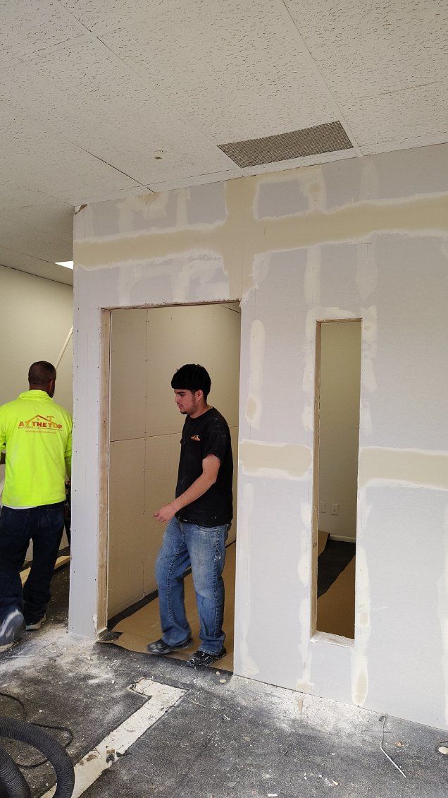 Men working on drywall in a room under construction. One walks through a doorway, the other in a reflective vest.