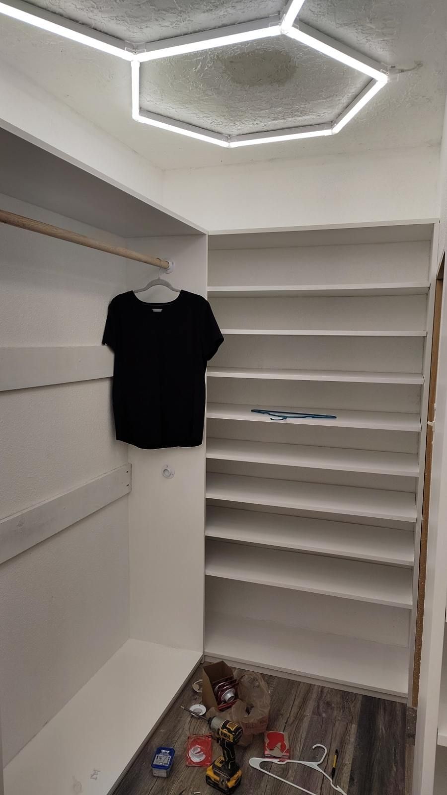 White closet interior with shelves and hanging rod. A black shirt hangs. Hexagon ceiling light.
