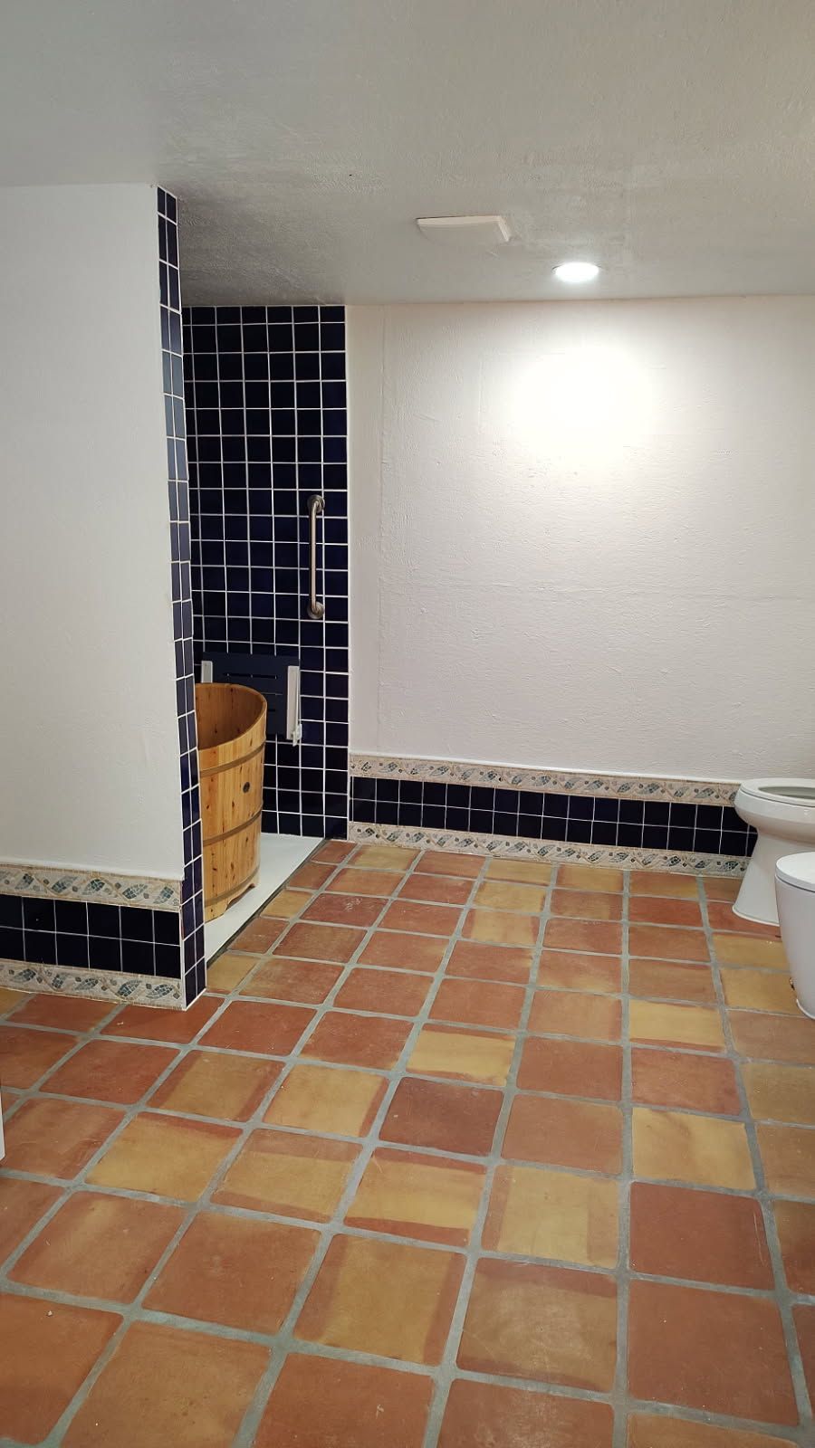 Bathroom with terra cotta tile floor, dark blue tile shower, wooden bucket, and white toilet.
