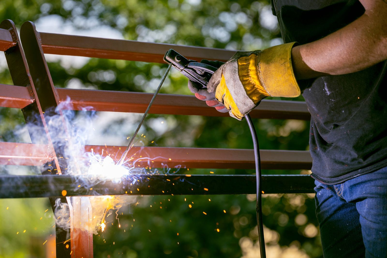 Welder in yellow gloves using a welding torch on metal beams outdoors, producing sparks and light.