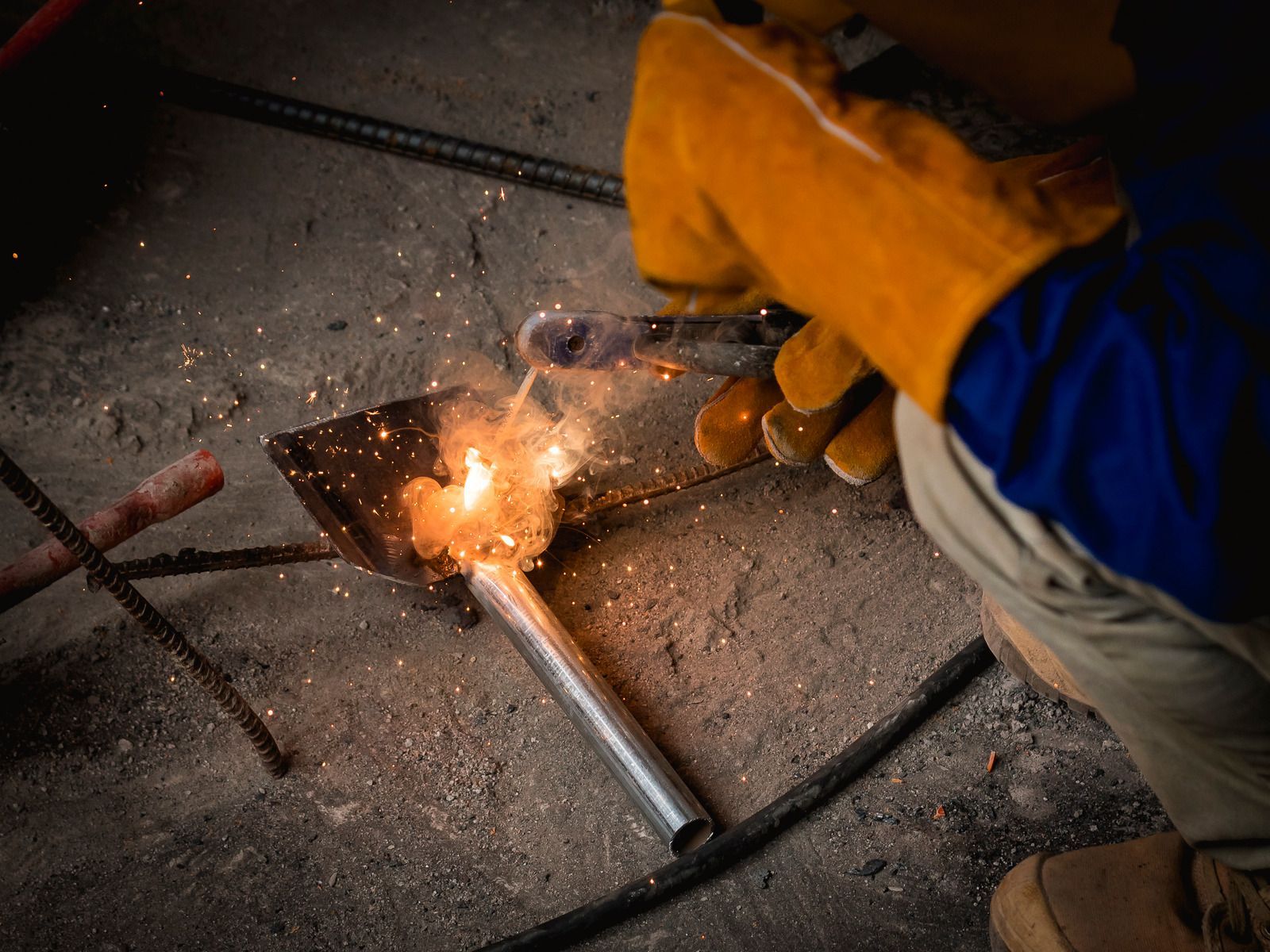 Welder wearing yellow gloves uses torch on metal tube, creating sparks in a workshop.