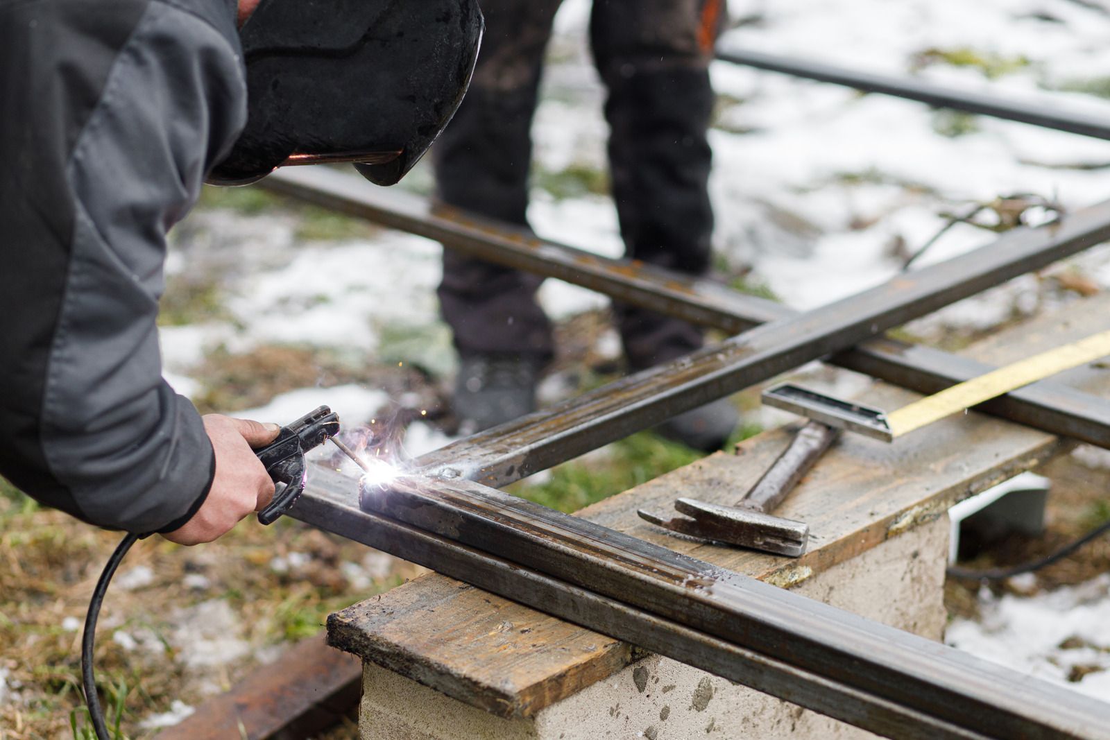 Welder in protective gear welding metal frame outdoors on snowy ground.