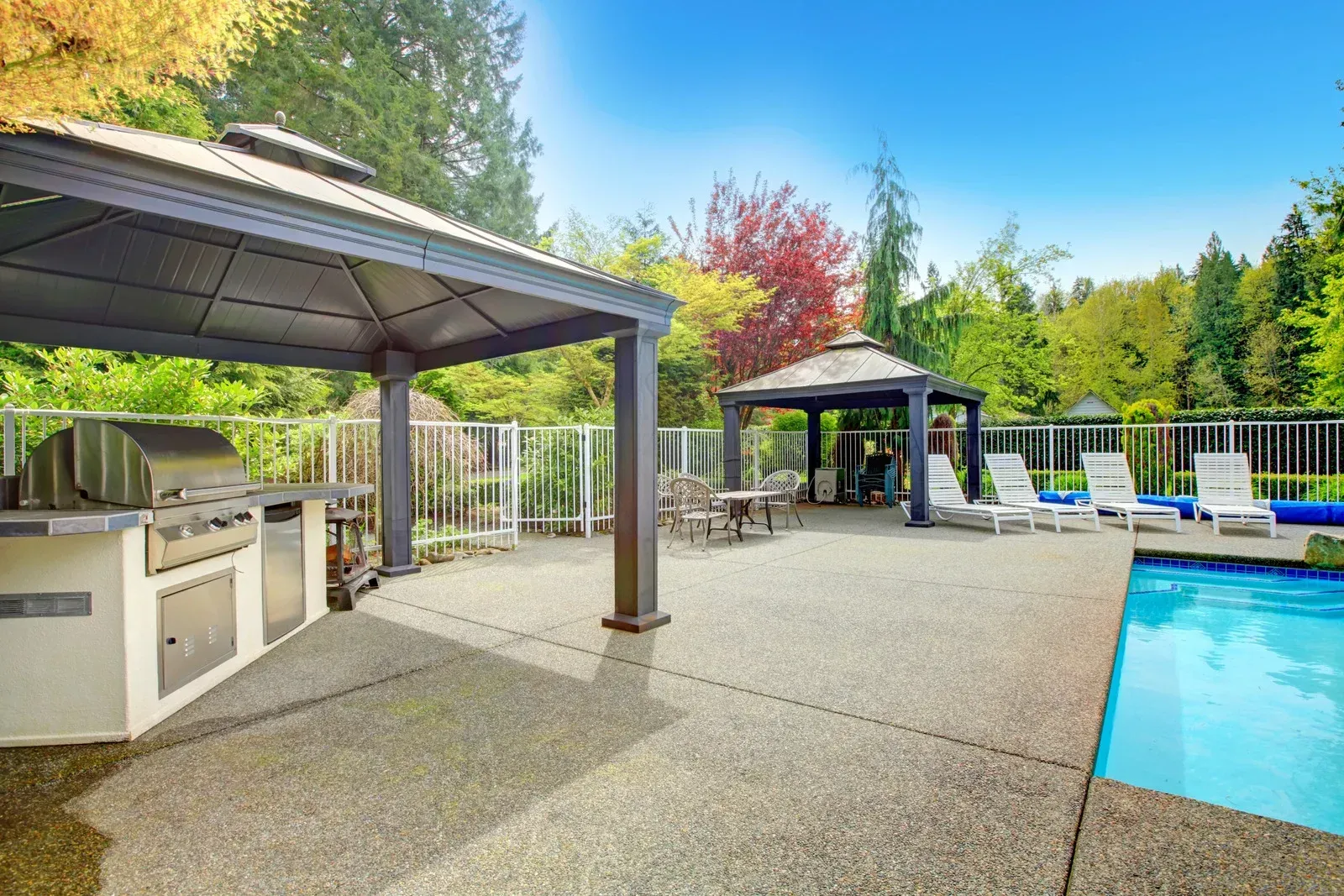 Patio with grill, two gazebos, pool, and lounge chairs under a blue sky and trees.