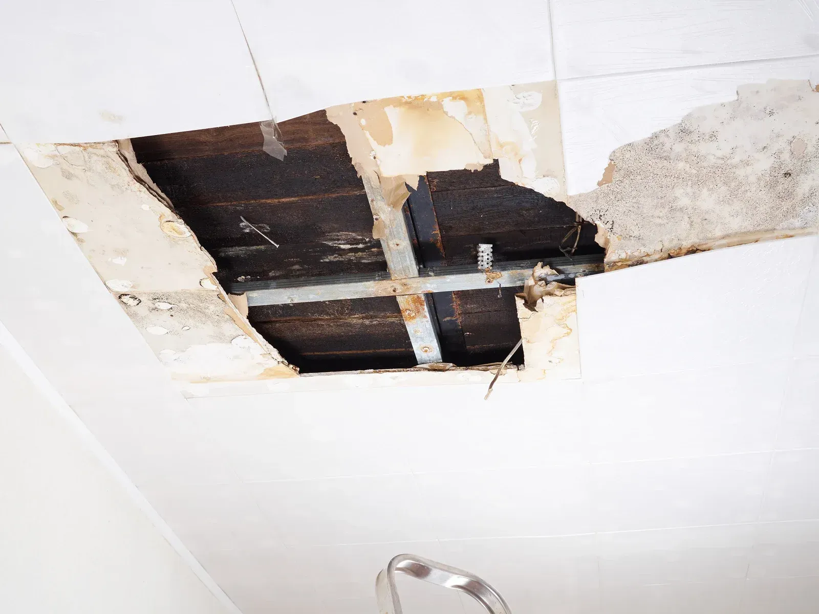 Hole in ceiling revealing wooden beams and metal supports, with damaged plaster and paint.