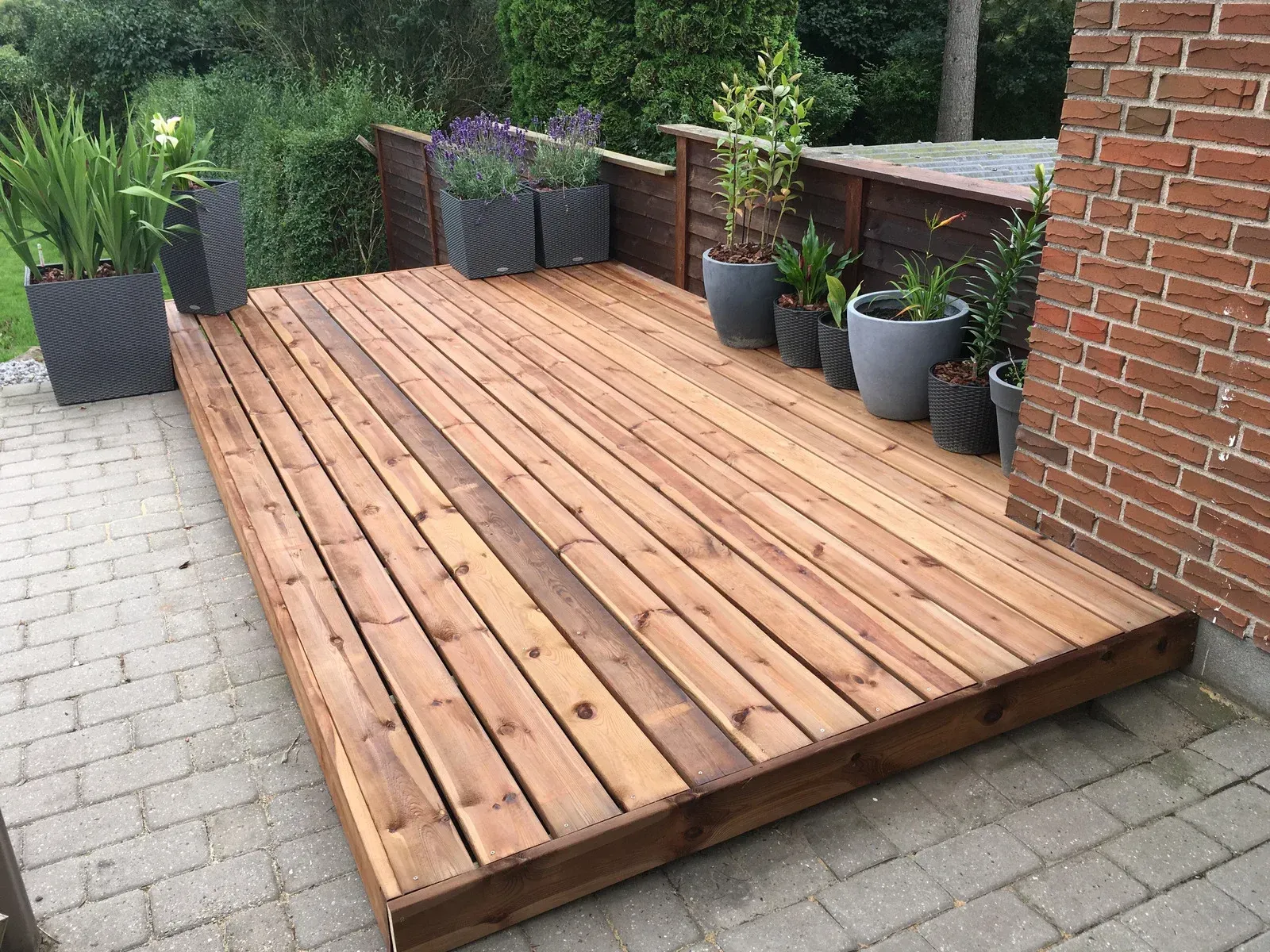 Wooden deck with potted plants against a brick wall and a hedge.