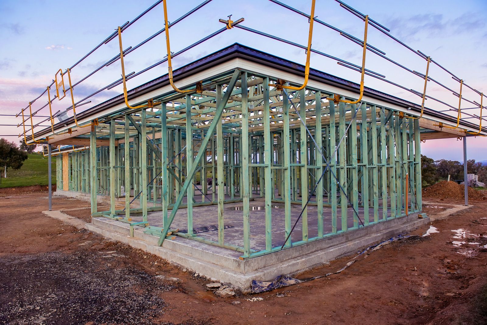 Construction of a house frame with green wood, concrete base, and scaffolding, set against a dusk sky.