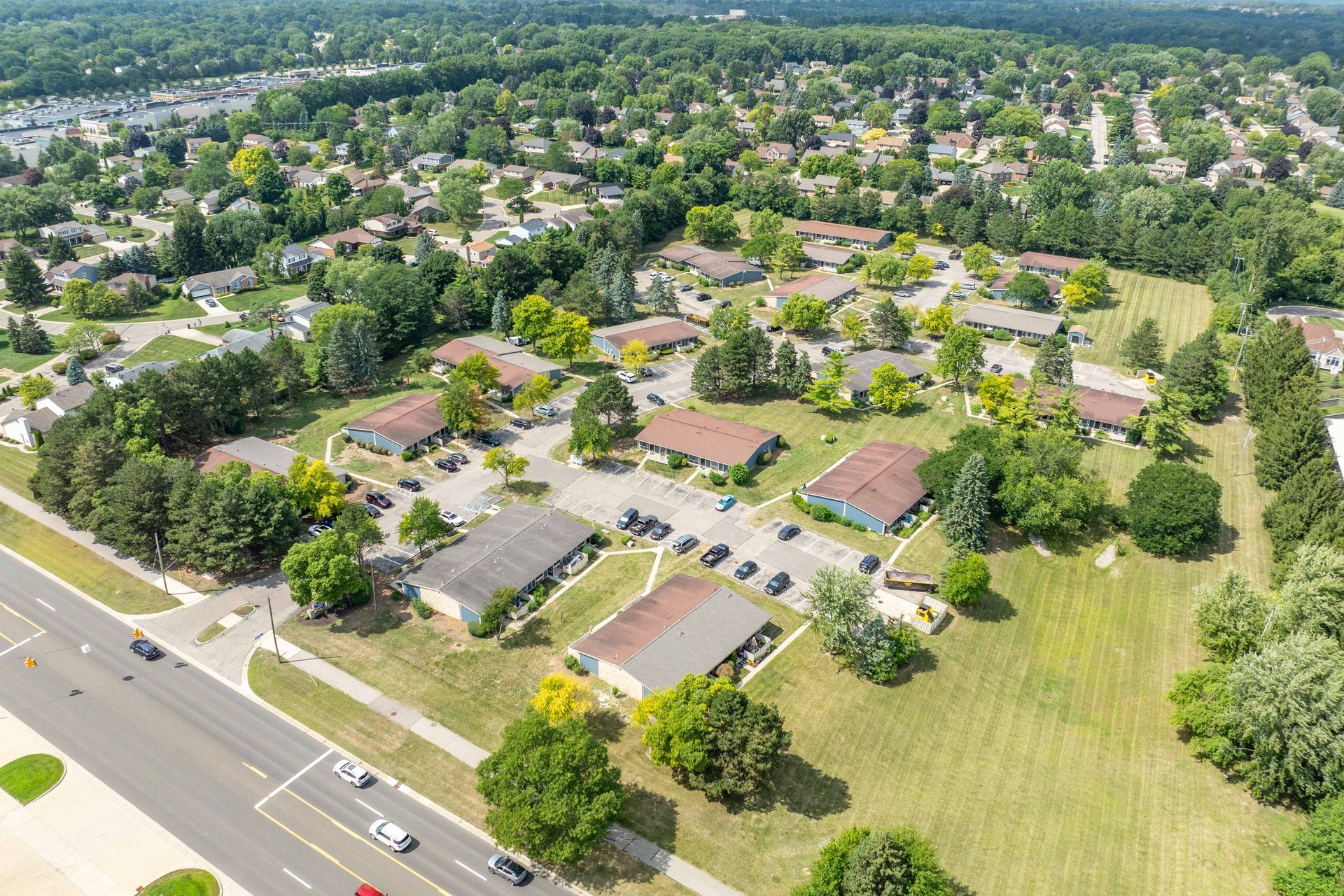 Aerial view of a residential neighborhood with low-rise buildings, green lawns, trees, and a road with cars.
