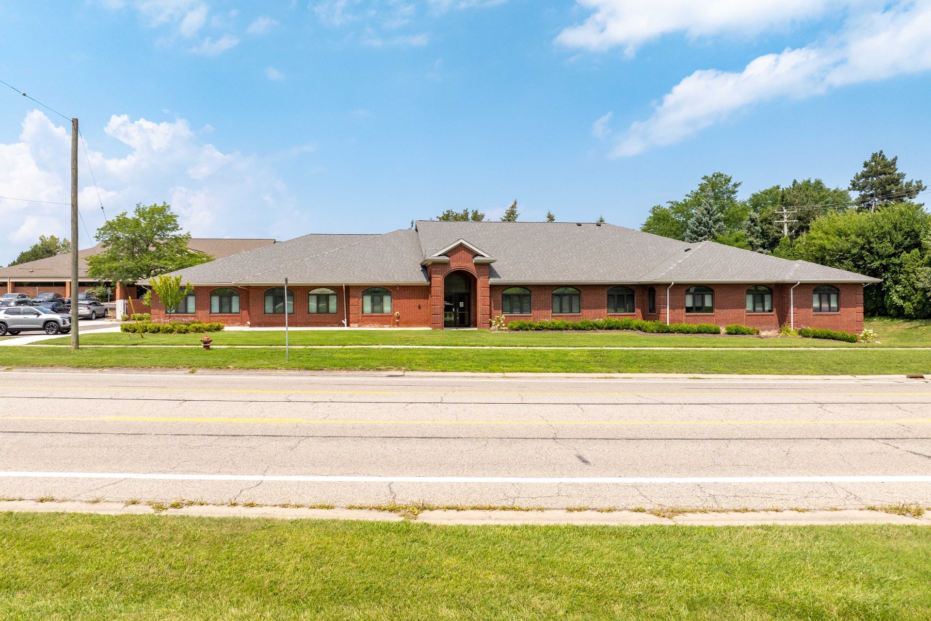 Red brick one-story building with a gray roof, set behind a road and green grass under a blue sky.