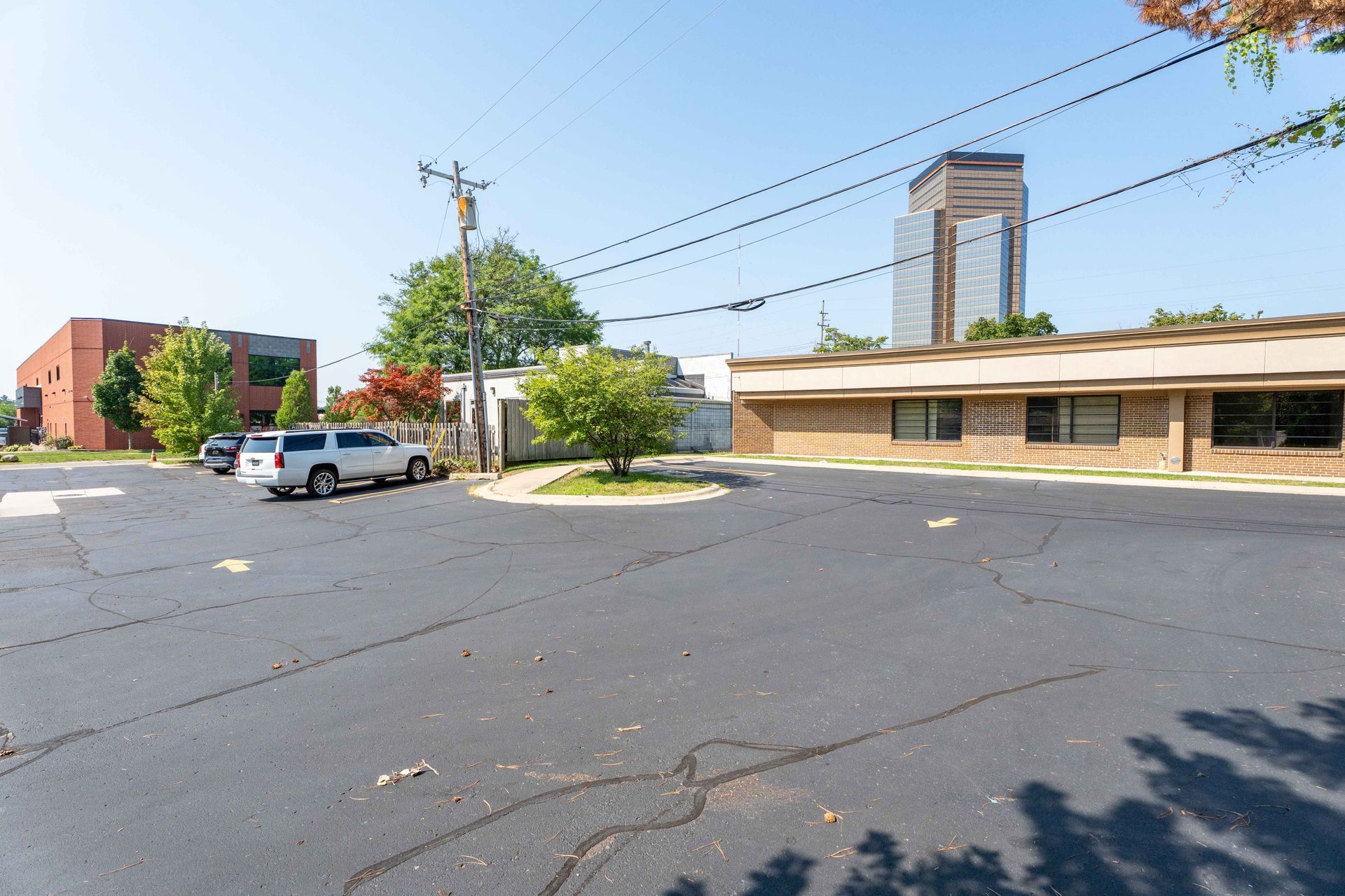 Paved parking area with a few vehicles, buildings, and a tall skyscraper in the distance under a blue sky.
