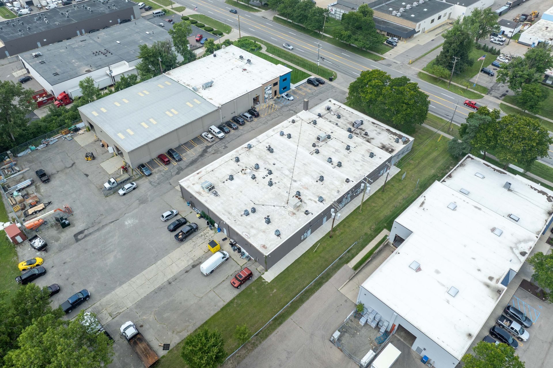Aerial view of several industrial buildings with cars parked outside. Green trees and a road visible.