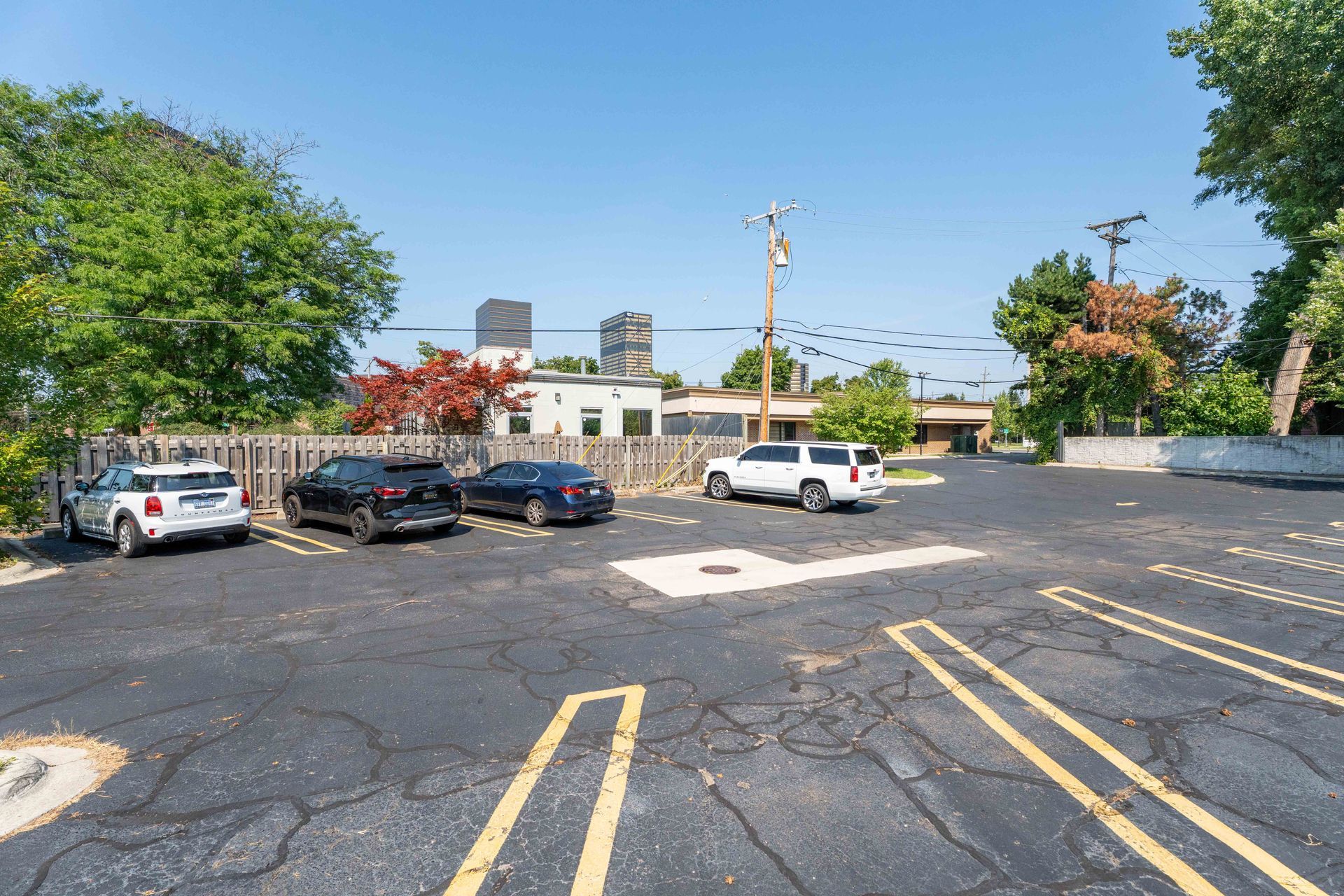 Parking lot with several vehicles on a sunny day; buildings and trees in the background.