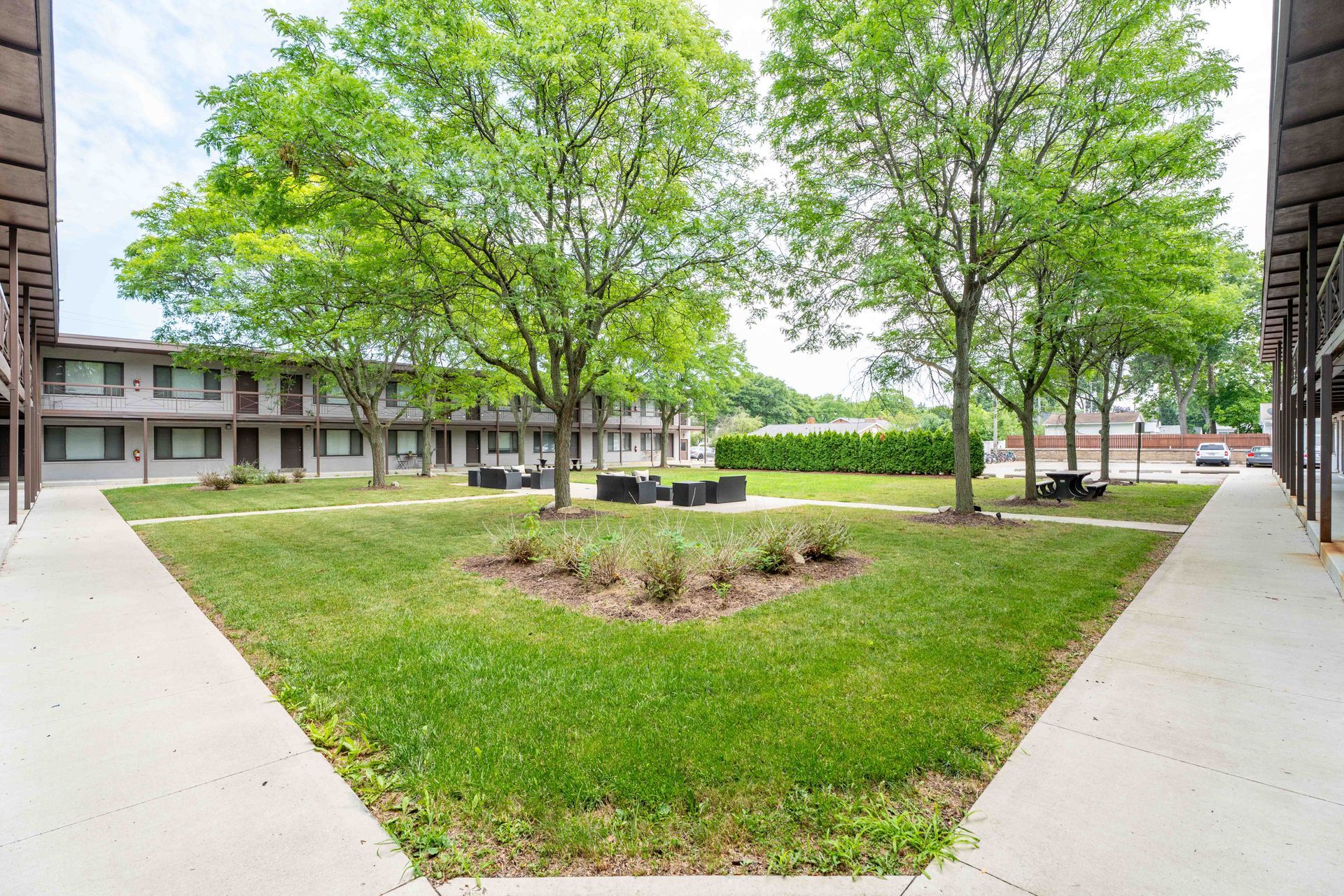 Courtyard with green grass, trees, and low seating in front of a motel building with brown siding and multiple levels.